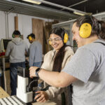 Zwei Mädchen mit Lärmschutzkopfhörern arbeiten gemeinsam an einer Handfräse in einer Werkstatt.