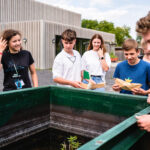 Eine Gruppe Jugendlicher steht lachend um einen Behälter mit Wasser mit einem Papierboot.