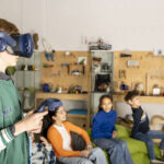 Ein Junge mit einer VR-Brille. Im Hintergrund sitzen zwei Mädchen und ein Junge auf Sitzsäcken.