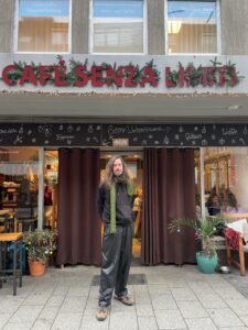 Marc steht vor dem Café Senza Limiti. Er ist ein Mann Mitte 30, hat lange Haare, einen Bart und trägt lockere schwarze Kleidung und einen grünen Schal.