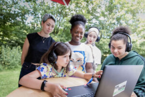 Auf dem Bild sind 5 Personen zu sehen, welche alle einen Laptop anschauen. Drei von diesen Personen tragen Kopfhörer.