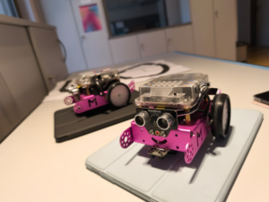 Zwei mbots sind auf einem Tisch abgebildet. Die pinken mbots sind ca. 12 cm lang und 8 cm breit und verfügen hinten über zwei Räder außen am Gestell. Vorne sind zwei Sensoren angebracht und mit der Einfräsung darunter sieht es wie ein lachendes Gesicht aus.