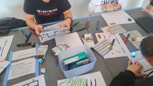Jugendliche sitzen gemeinsam am Tisch und interagieren mit den Ozobots. Es sind verschiedene Parkours auf Blätter aufgezeichnet. Die Ozobots sind kugelförmig mit einer flachen Seite unten, wo die kleinen Räder und die Sensoren angebracht sind. Die Ozobots haben einen Durchmesser von ca. 4 cm und können blinken.