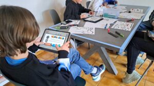 Hier werden Jugendliche abgebildet, die mit Tablets an einem Tisch sitzen und "Draw your game" spielen. Auf dem Tisch liegen Blätter, auf denen verschiedene Parkours gezeichnet wurden.