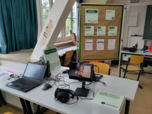 Auf der linken Seite im Vordergrund sind zwei Mitmachaktionen abgebildet. Bei einer Mitmachaktion sind Laptops mit der Browser-Erweiterung "Disability Web Simulator" vorbereitet. Bei der anderen Mitmachaktion ist auf einem Tablet ein Video vorbereitet, in dem gezeigt wird, wie einer stummen Person von einer Stimmpatin die Stimme geliehen wird.
