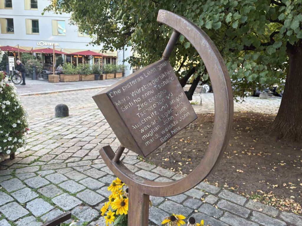 Mahnmal für die im Nationalsozialismus verfolgten und ermordeten Sinti und Roma auf dem Paradeplatz in Würzburg. Die rostfarbene Metallskulptur besteht aus einem offenen Ring mit einer schräg angebrachten Gedenktafel. Am Fuß des Mahnmals liegen gelbe Blumen.